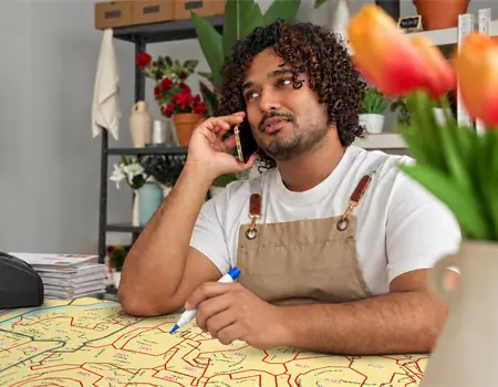 A man on the phone in a flower shop, marker in hand, leaning over a yellow carrier route map outlined in red spread across the counter.