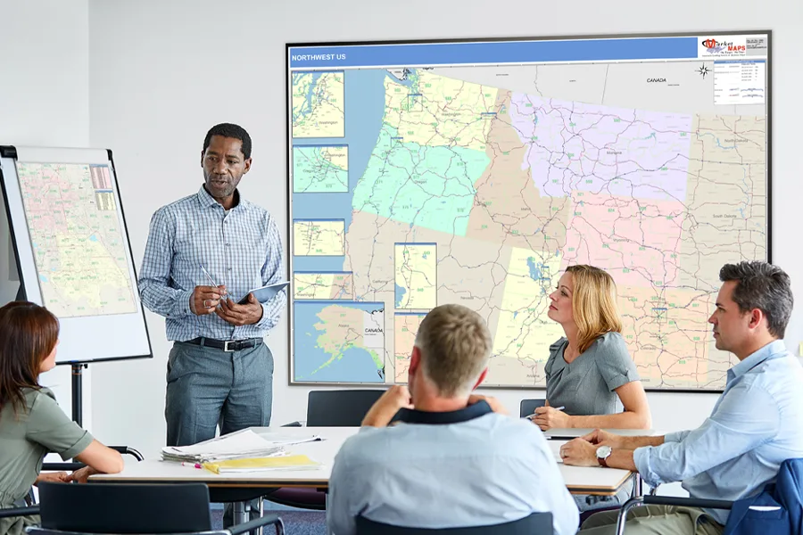 A presenter holding a tablet in front of a Northwest US wall map, its states colour-coded into territories, while four colleagues listen around a conference table.
